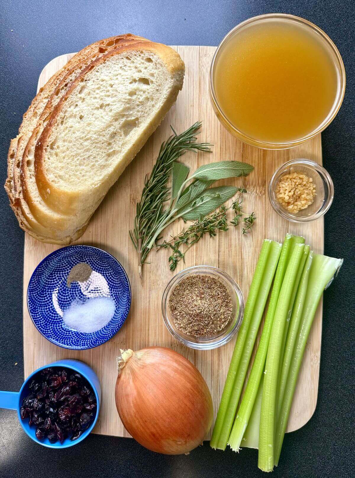 Overhead view of ingredients for vegan sourdough stuffing, including sourdough bread, celery, onion, herbs, dried cranberries, and vegetable broth.